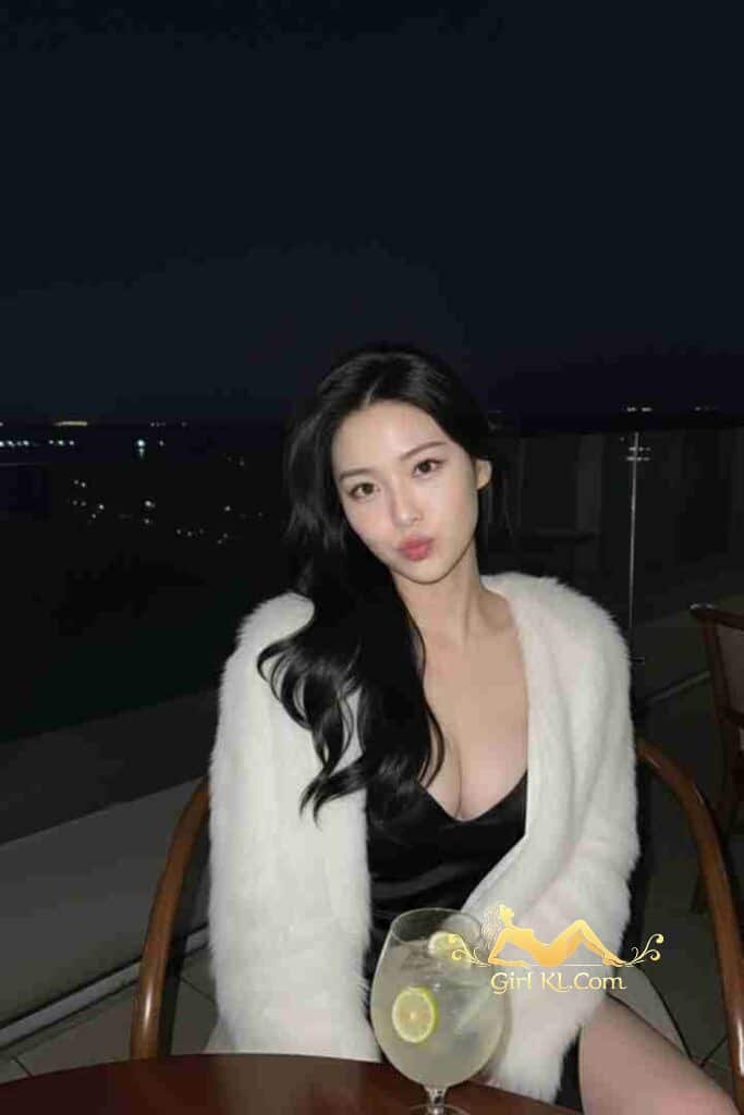 Elegant Asian woman in black dress & white fur jacket on balcony at night. VIP KL Escort Qi Qi.