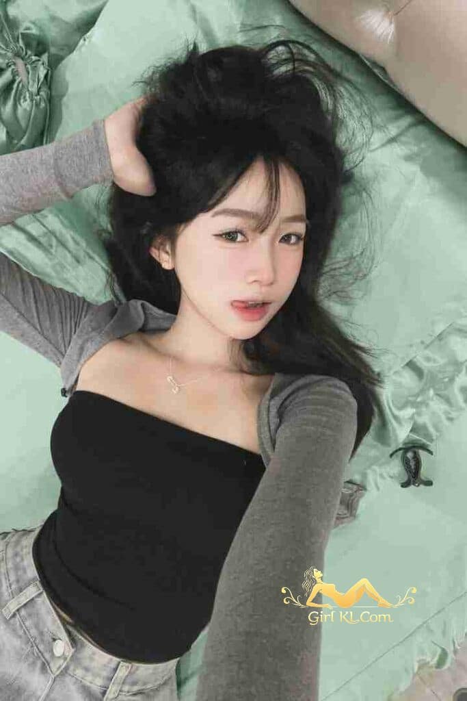 Attractive Asian woman with dark hair laying on a bed with a grey top and jeans.