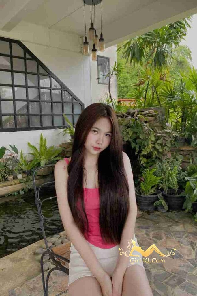 Stunning Thai woman with long hair sitting outdoors, pink top, white shorts.