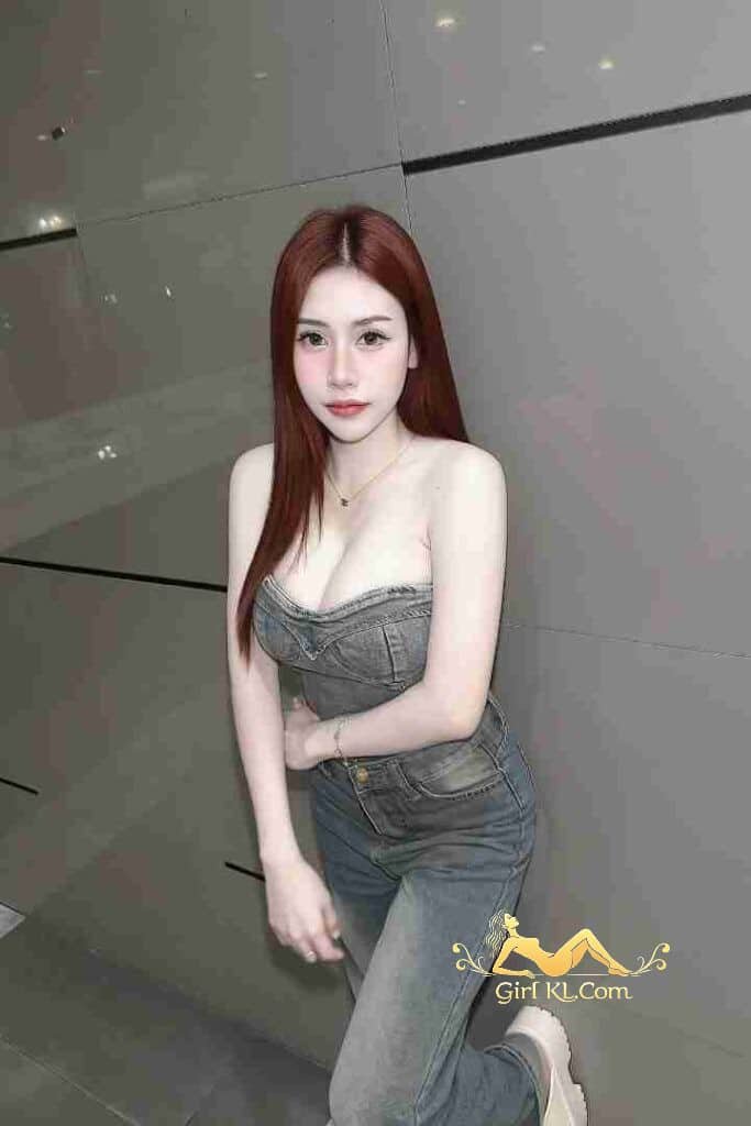 Stunning Thai woman with red hair in denim top and jeans posing against a wall.