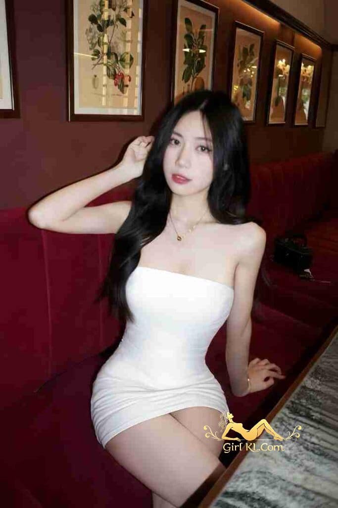 Vietnamese woman in white dress on red couch. Black hair, necklace. C Cup beauty, Kuala Lumpur.
